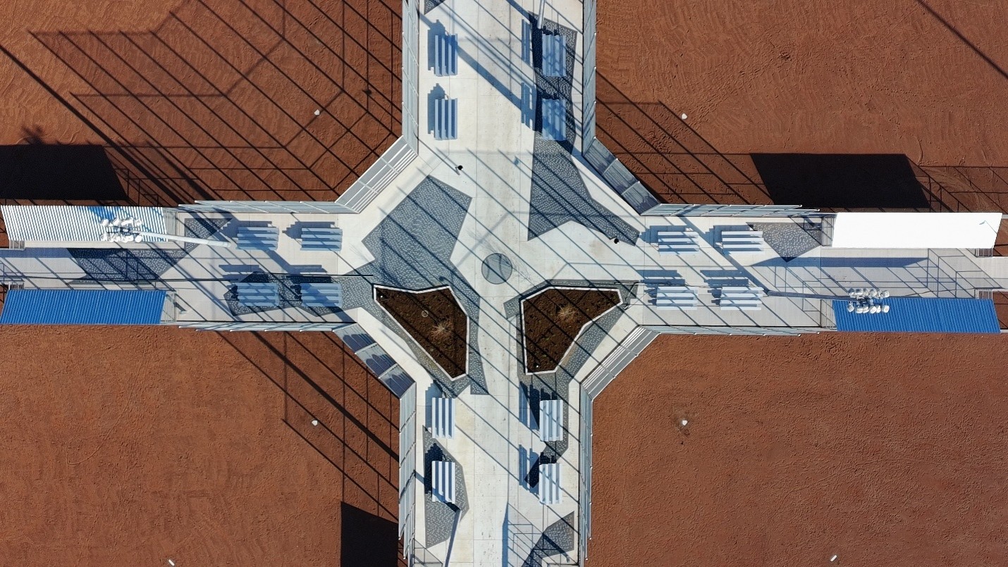 Overhead aerial view of the central hub connecting the reddish-brown infields of four baseball diamonds. Concrete walkways form a cross-shaped plaza with seating, fencing, planting beds and blue-roofed dugouts arranged symmetrically around the centre.