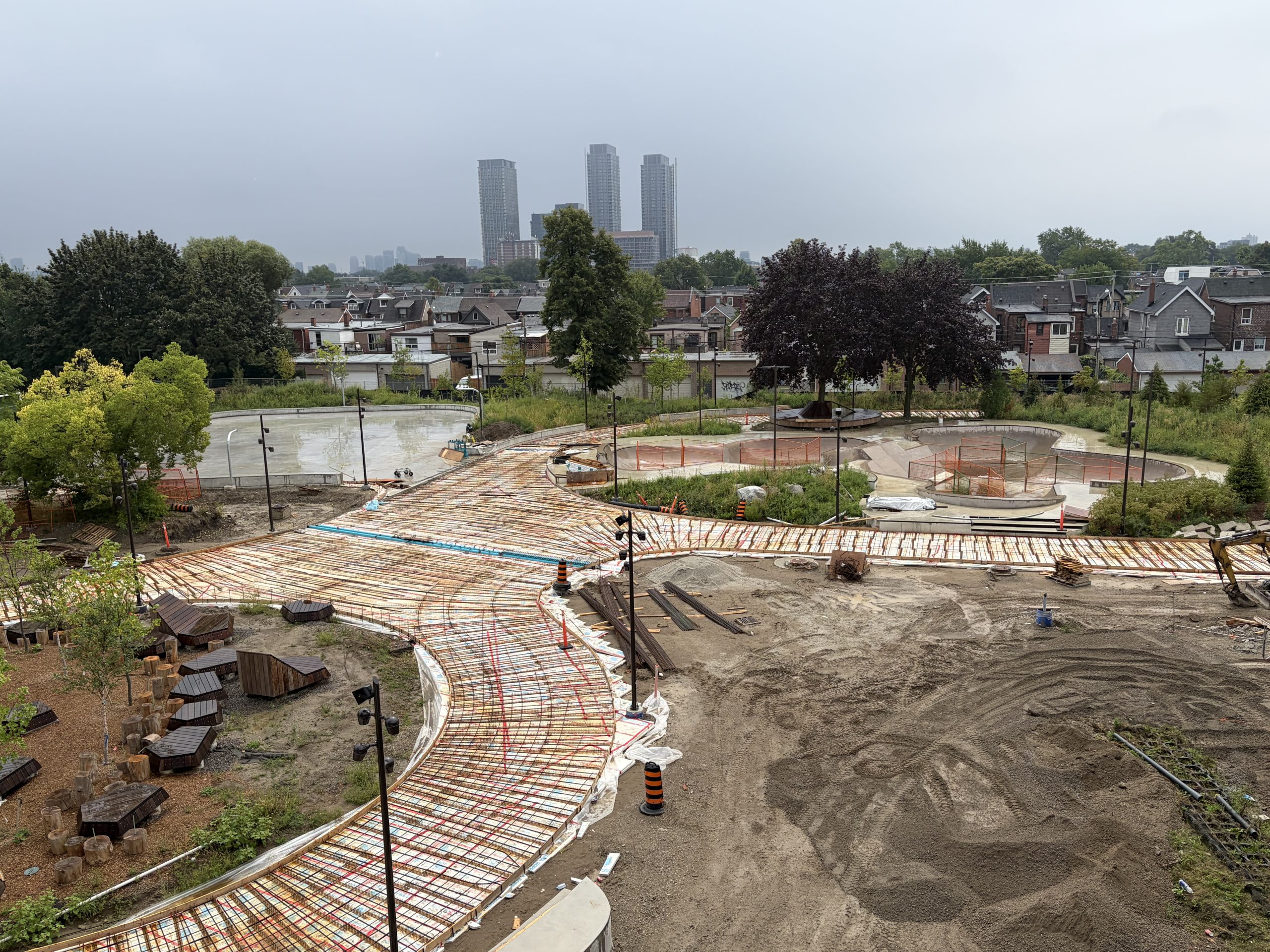 Aerial view of a construction site, showing steel mesh installations atop white plastic coverings that form the course of a wide promenade in the shape of an eight. One loop surrounds a landscaped area with new tree plantings, decorative tree stumps and wooden deck furnishings. Another loop surrounds a skate park with concrete bowls. Nearby, construction materials are scattered on dirt. Residential houses and taller buildings in the distance line the background.