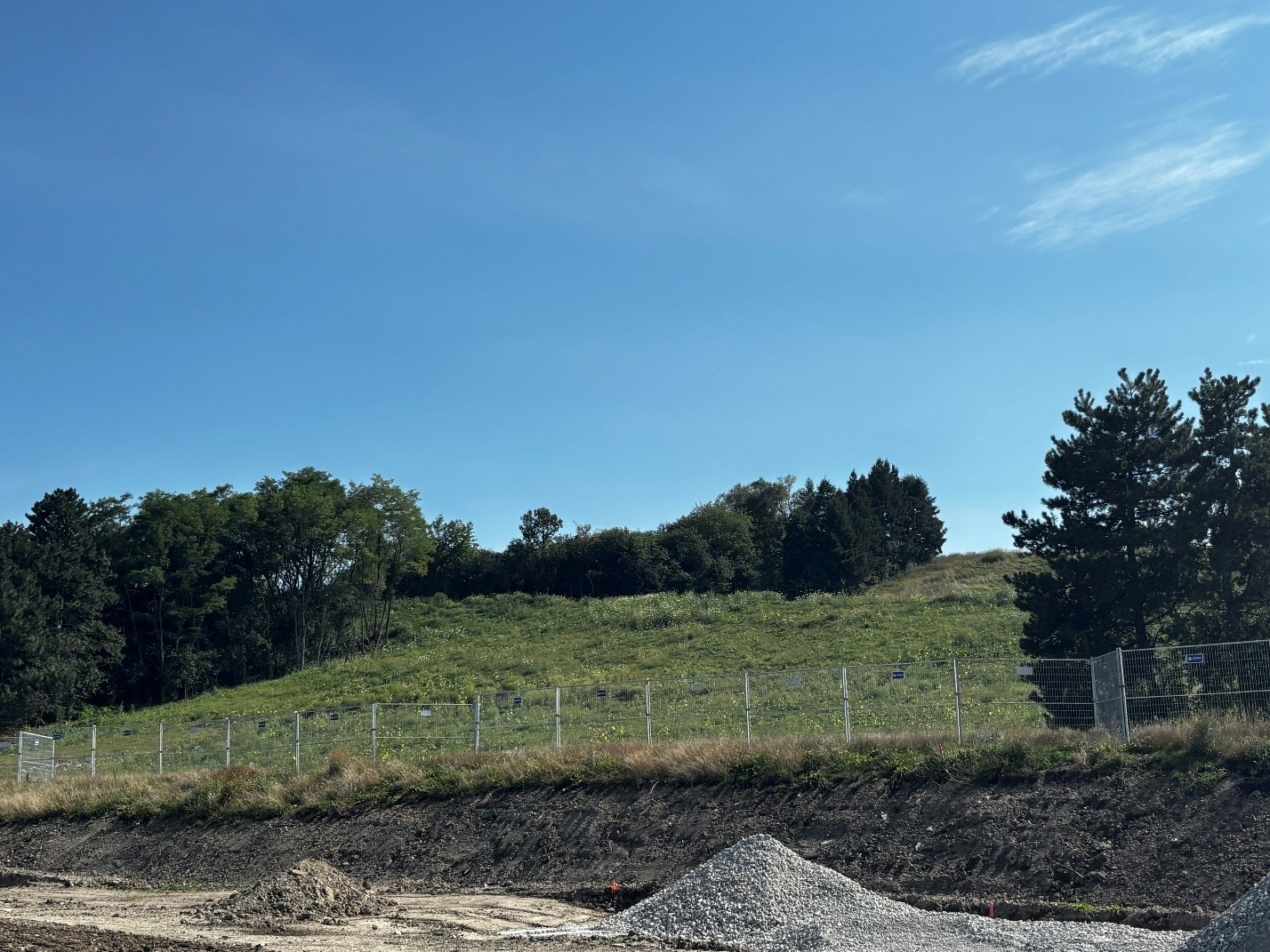 Grassy hillside with a temporary construction fence running along the slope. Mature trees line the top and sides of the hill, while piles of gravel and exposed soil are visible in the foreground.