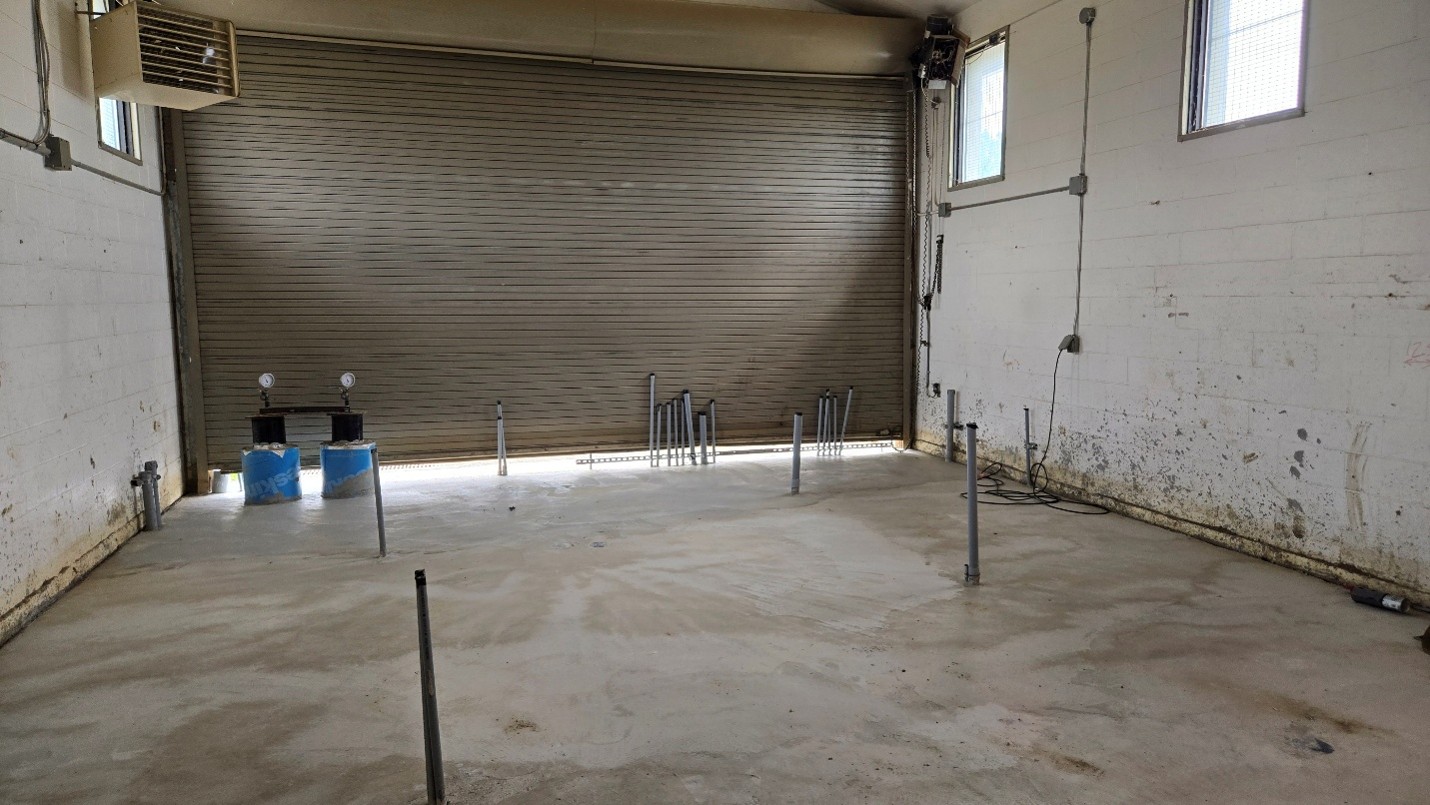 Interior view of a room with a concrete floor, block walls and a metal roll-up door. Multiple pipes extend vertically from the floor. Exposed wiring and high windows line the space.