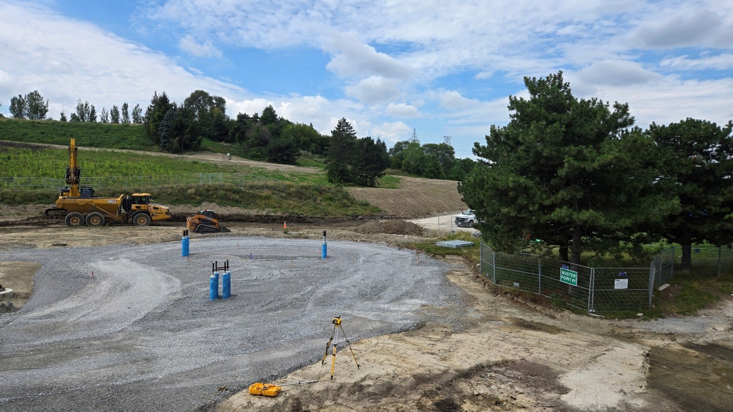 Construction site in a park setting, featuring a circular gravel area and several blue utility pipes extending from the ground. An excavator, dump truck and compact track loader are positioned along the left side of the site.
