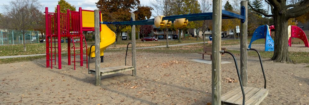 Playground with a red climbing structure and yellow spiral slide, an overhead track ride, and a blue climbing wall, set on a sand surface with mature trees in the background.