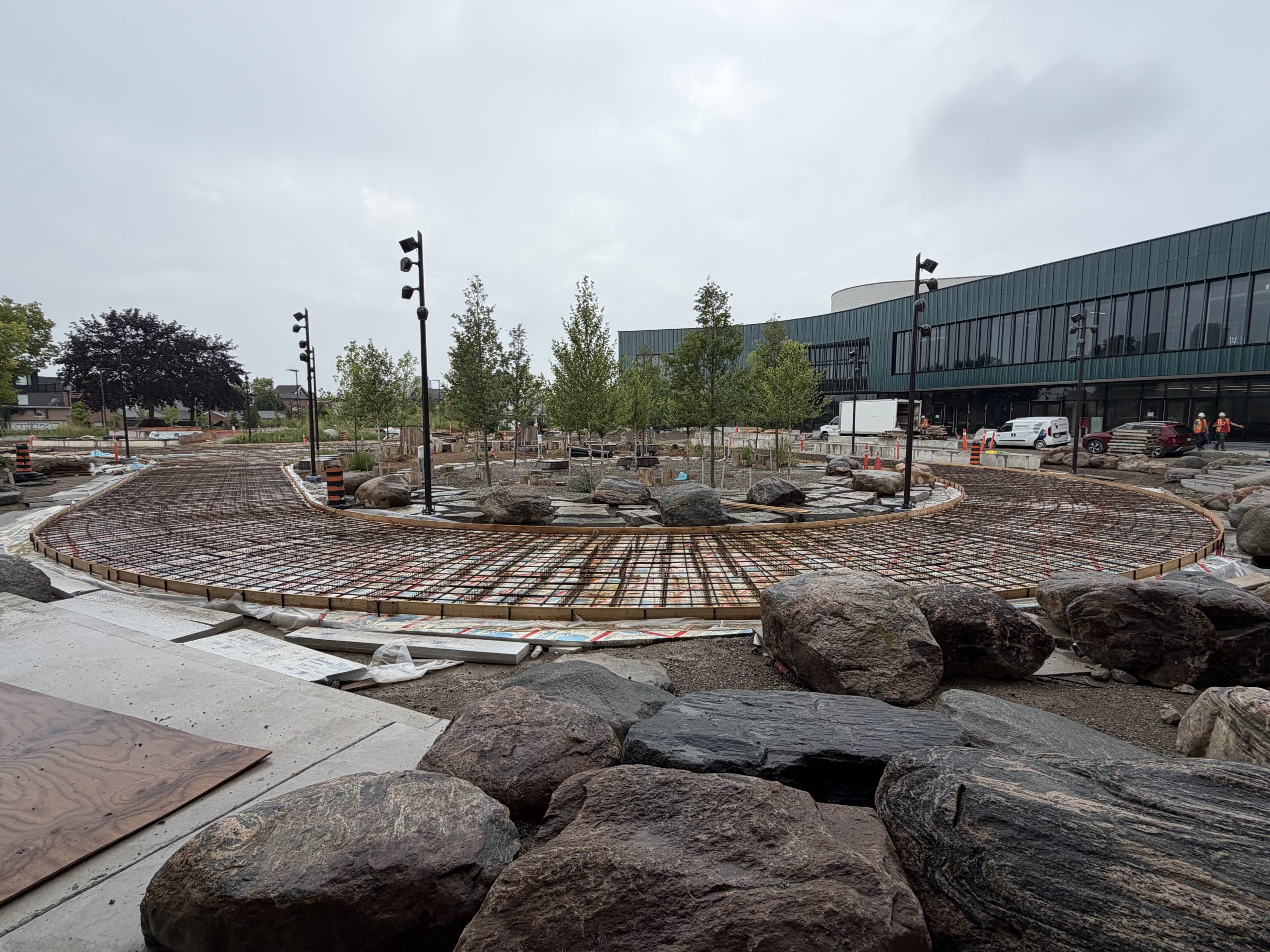 A circular plaza under construction beside a modern building with teal cladding. Steel mesh installations form the course of a pathway that curves around a central landscaped area with newly planted trees, decorative boulders and light poles. More decorative boulders are visible in the foreground. Construction materials and workers are seen throughout the site.