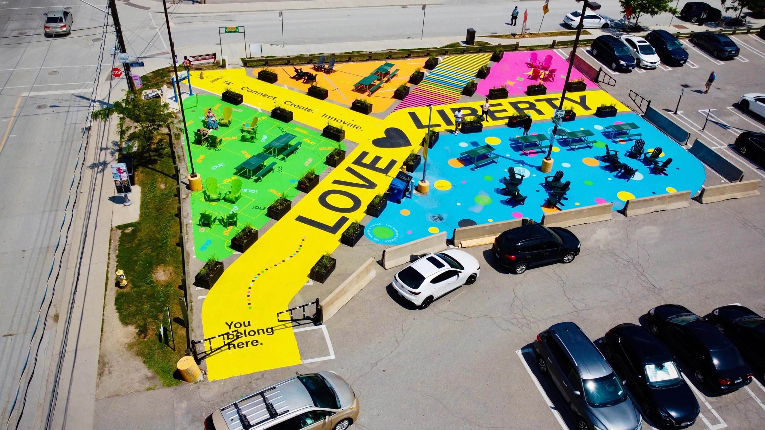 Aerial view of the Park in Progress space located at the northeast corner of the existing parking lot site (Hanna Avenue and Liberty Street), showing the brightly coloured mural painted in green, yellow, blue, orange and pink, with large black lettering that reads “LOVE LIBERTY”. Tables, seating areas, and planters are distributed throughout the site, which is surrounded by streets and parking lot.