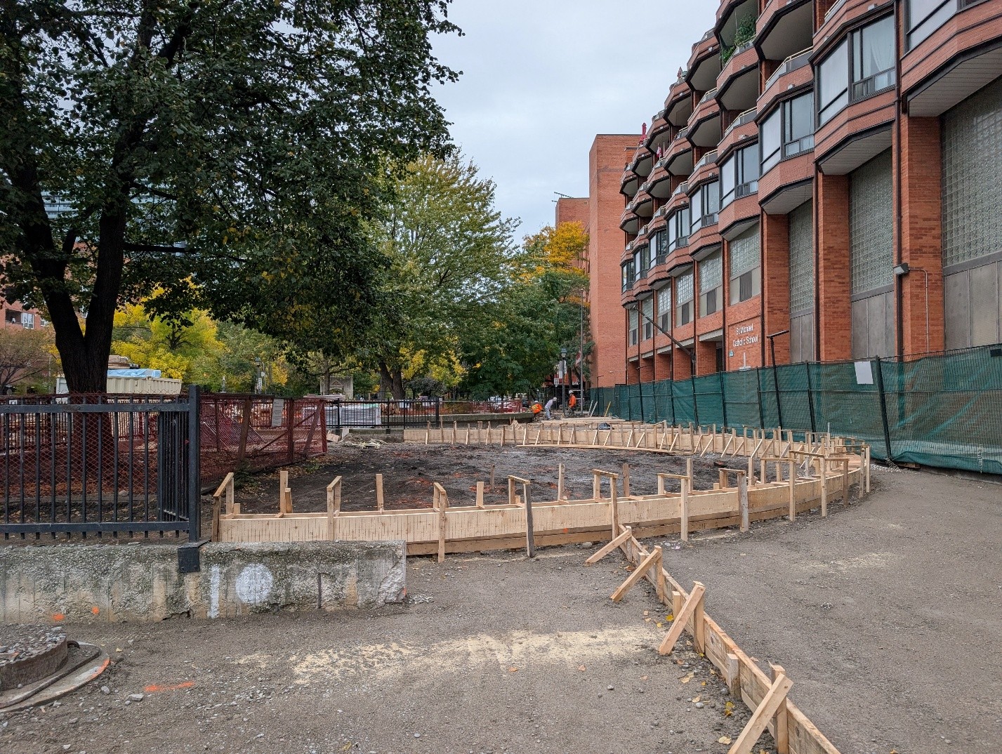 Construction site at Block 2 with wooden concrete forms outlining a paved pathway or area of the park. Mature trees surround the site on one side, while green construction fencing runs along the other side, abutting a brick building.