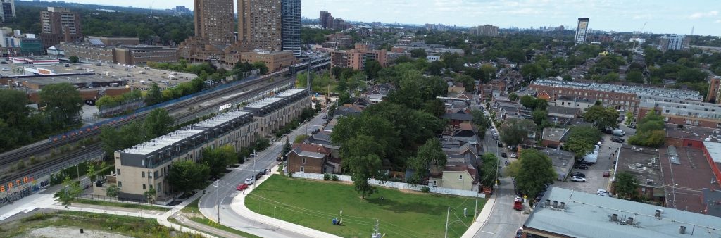 Aerial view of a vacant development site beside a tree-lined residential neighbourhood, with rail tracks, streets, and mid- and high-rise buildings nearby.
