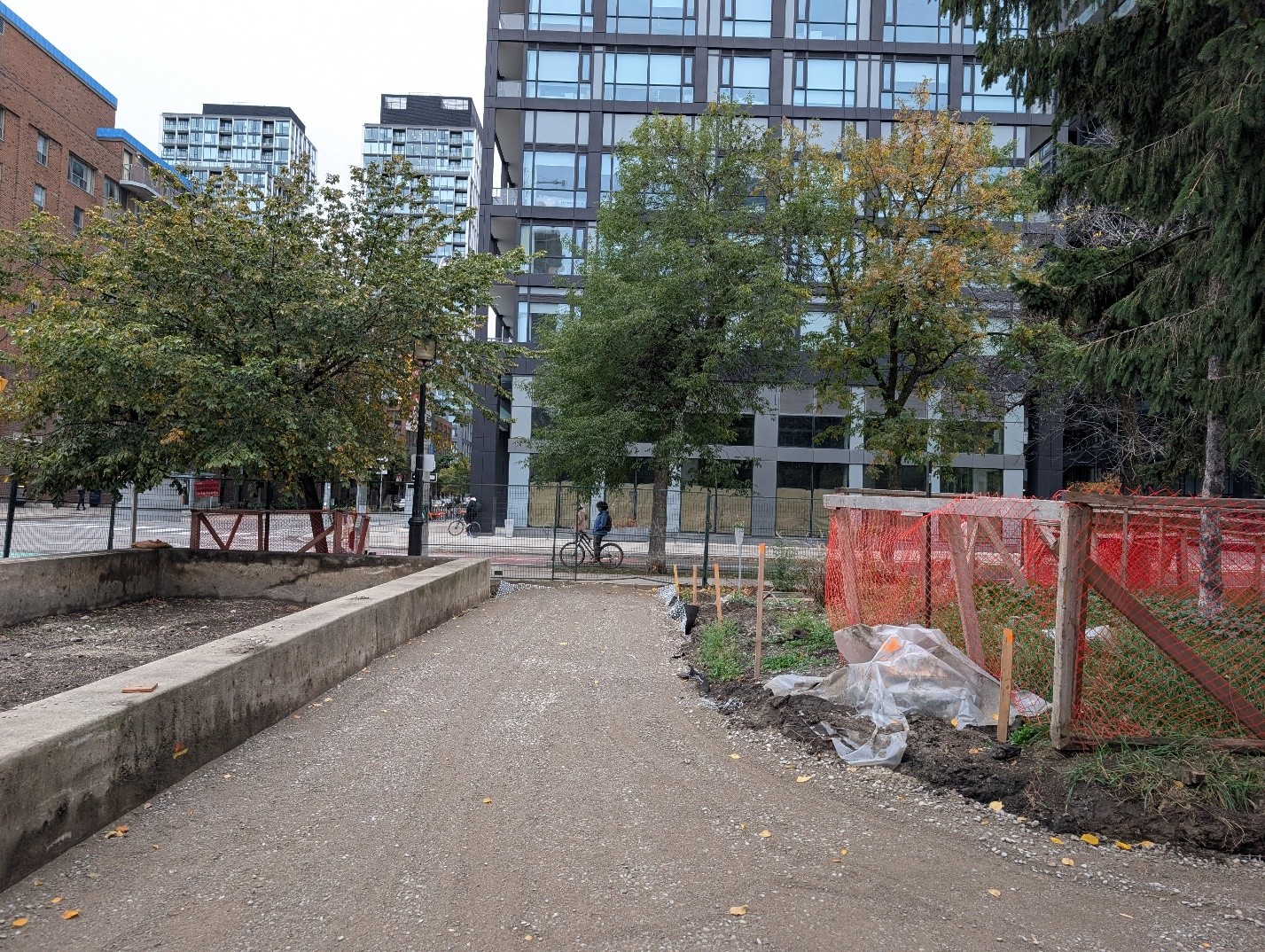 Construction site at Block 5, with a gravel pathway bordered by low concrete walls and temporary safety fencing. Plantings and exposed soil line the path, with mature and residential buildings in the background.