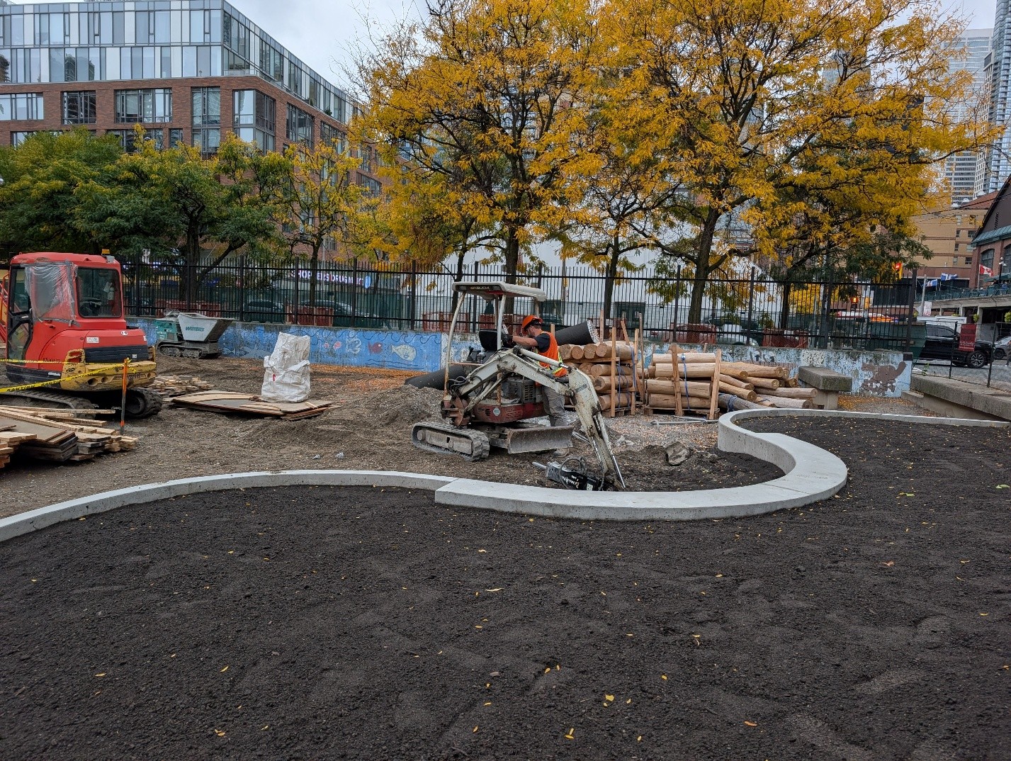Construction site at Block 2 with construction machines near stacks of timber. A large, curved area in the foreground is filled with soil and bordered by concrete. Mature trees and multi-storey buildings are visible in the background.