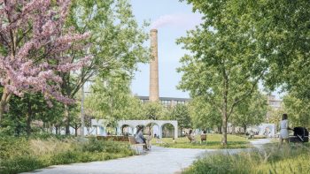 Eye-level view along a concrete paver path, spring blossoms flowering at the left and fresh green canopy overhead, billowing planting beds running alongside the path, the low pavilion crossing the middle distance and the heritage brick chimney rising behind.