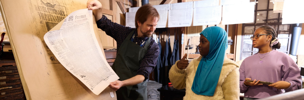 Historic interpreter shows a broadsheet to two students in a historic print shop