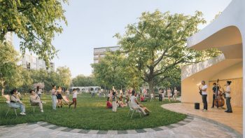 Eye level view of a curved stage with an acoustic band playing underneath and a grass lawn in front with people lounging and watching the performance. In the background are a few local buildings that surround the park, some trees and the low pavilion. In the foreground is a walkway with stone paving.