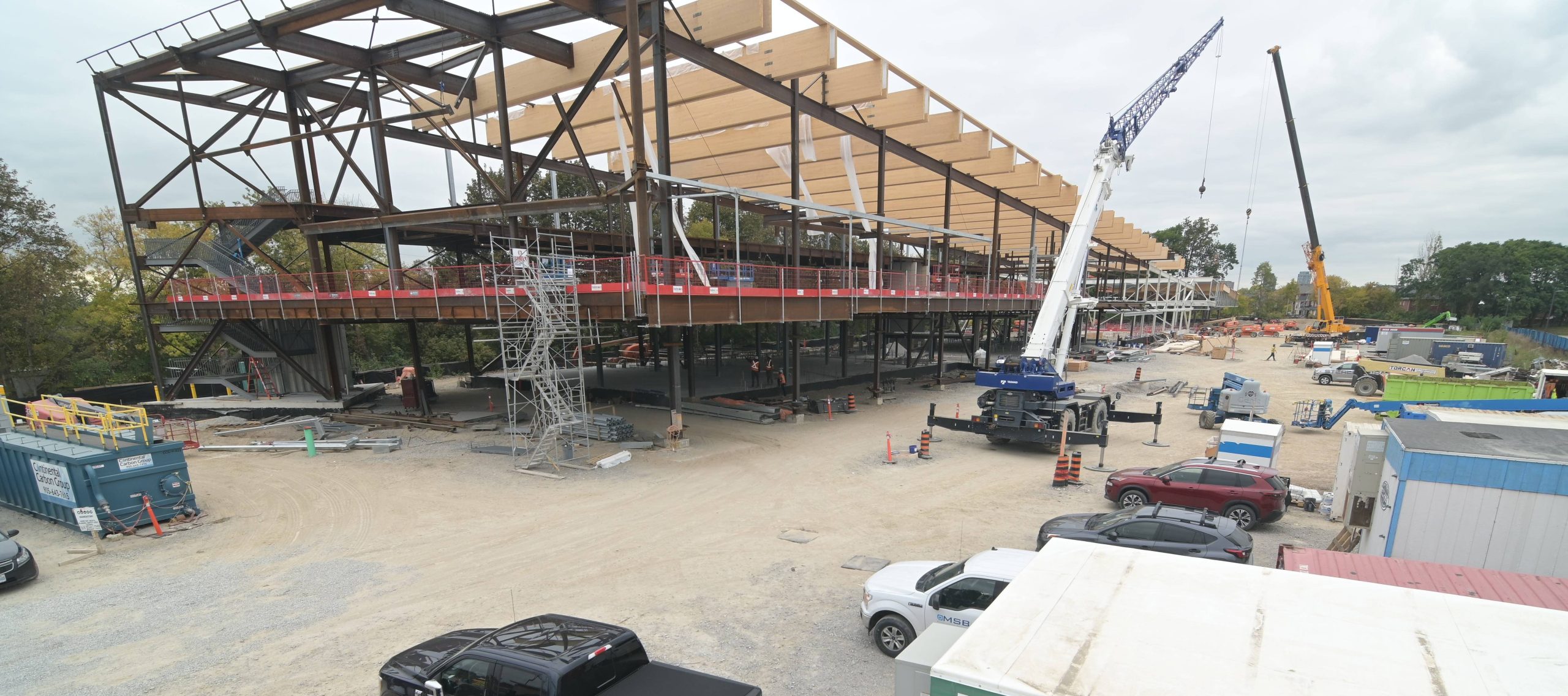 Side view of the new community recreation centre under construction, showing the structural steel frame and timber installation in progress. The large rectangular building frame sits on a dirt and gravel lot. Workers are visible around the site, while cranes lift materials for the building. Parked cars and shipping containers are also present on site.