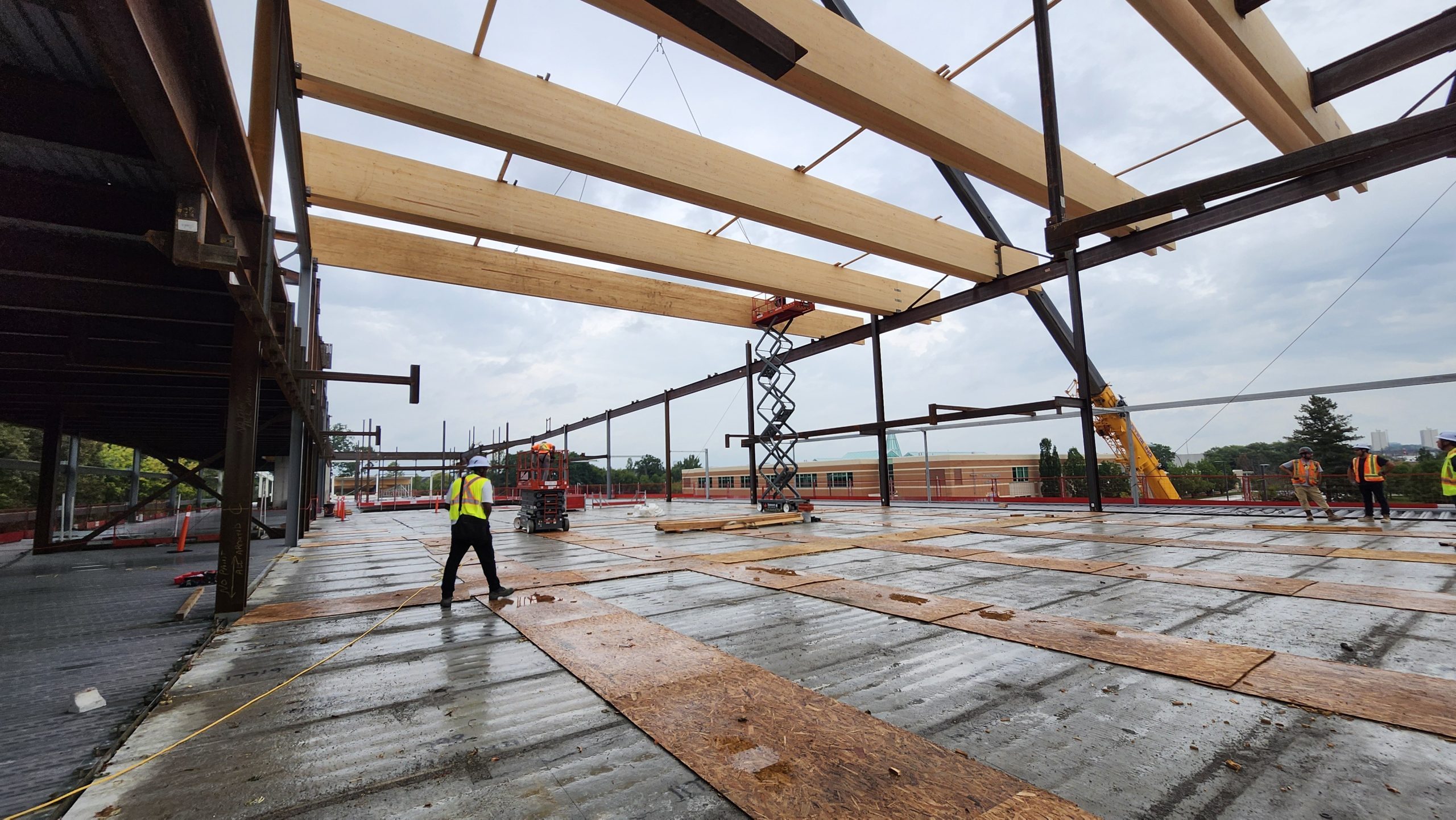 View of the new community recreation centre under construction, showing the mass timber installation in progress over the second storey. Thick wooden slabs are positioned horizontally across the structural steel building frame. Thin wood boards are arranged in a grid pattern on the ground. Workers, lifts and a crane are visible on site.