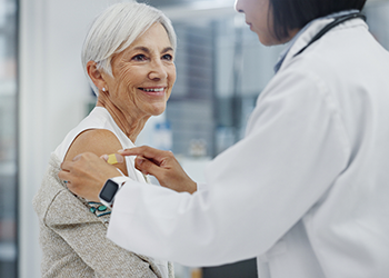 A healthcare worker applies a small adhesive bandage to the upper arm of an older adult during a clinic visit. The older adult is seated and wearing a light-colored sweater while the healthcare worker in a white coat stands beside them. The image focuses on post-vaccination care in a healthcare setting.