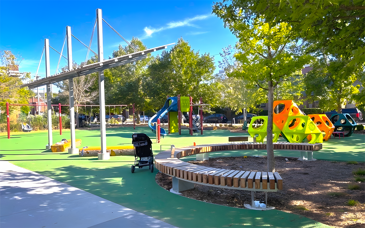 Zoomed out view of the new Wychwood Barns Park playground, showing the shade structure and a swing set on the left, a main play structure with two slides in the middle and play cubes to the right, a circular bench in front and trees in the background.