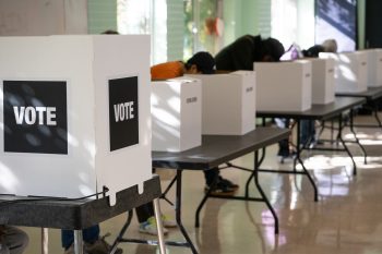 Voters marking ballots at a row of privacy screens inside a polling location.