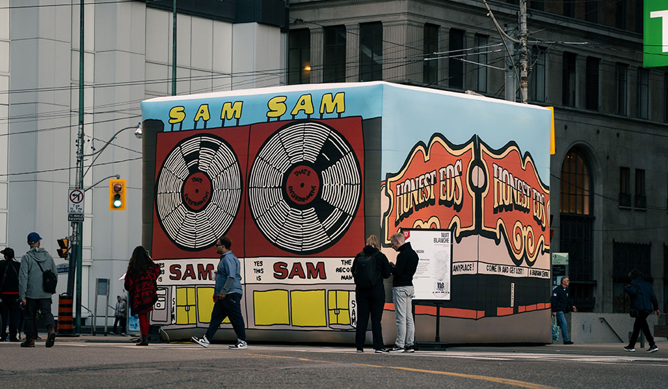 Large cube in downtown intersection with illustrations on the sides. Left visible side shows Sam the Record Man and right visible side shows Honest Eds. Some people walk in front.