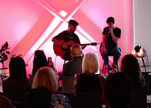 A singer and guitar player perform for an audience in front of a neon pink wall.