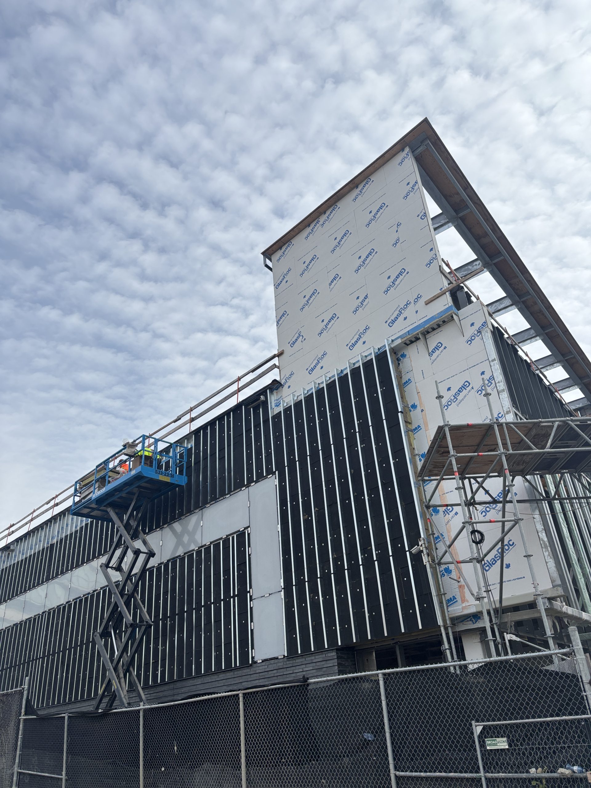 The community centre exterior under construction, with sheathing and black wall panels being installed. Scaffolding surrounds the building, and a scissor lift with workers is visible. A chain-link construction fence runs across the foreground at the base of the structure.