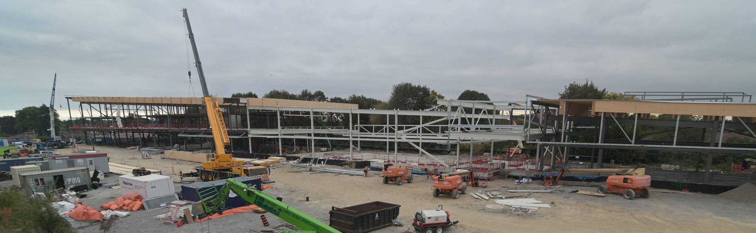Wide view of the construction site for the new community recreation centre. Structural steel forms the frame of a large rectangular two-storey building. Wood panel installations are underway on the roof. The site includes large cranes, trucks, shipping containers and other equipment and materials scattered around.