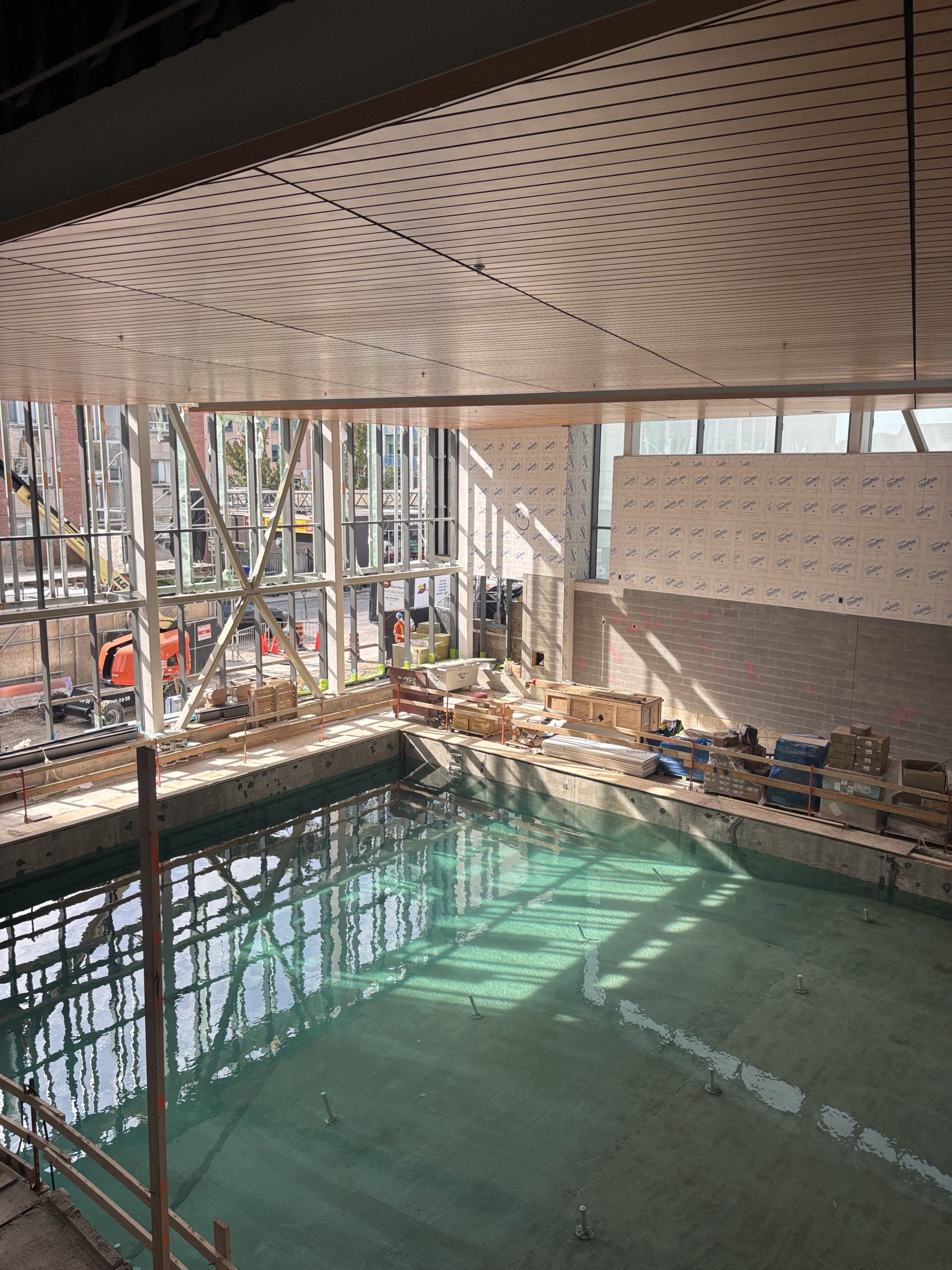 The indoor swimming pool in progress at the community centre, with floor-to-ceiling windows on one side that let in natural light. The adjacent wall is partially finished with grey brick. Most of the space is taken up by the pool, which has been filled with water. Crates and piles of construction material are scattered around.