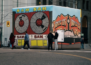 Large cube in downtown intersection with illustrations on the sides. Left visible side shows Sam the Record Man and right visible side shows Honest Eds. Some people walk in front.