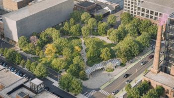 Aerial view of the completed park with its tree canopy grown in, cloud-like lawns of overlapping rounded lobes and paved paths winding between mature trees, the entry pavilion and its solar canopy along the street at the lower right and the heritage brick chimney at the right edge.