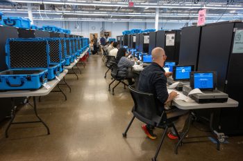 Toronto Elections workers conduct logic & accuracy testing on tabulators at individual stations inside a large warehouse.