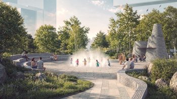View of the civic plaza with children playing within the misting feature, people sitting on the amphitheatre terraces and seatwalls around, and the washroom pavilion to the right in the background.