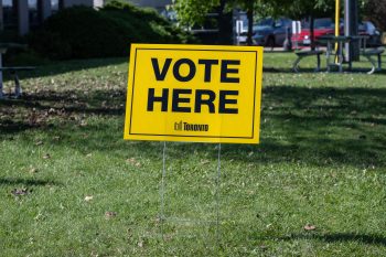 Yellow lawn sign reading “VOTE HERE” with the City of Toronto logo, placed on grass outside a voting place.