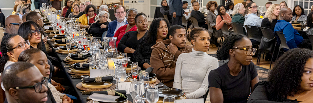 a long banquet table set for a meal. People seated at each place are watching a presentation.
