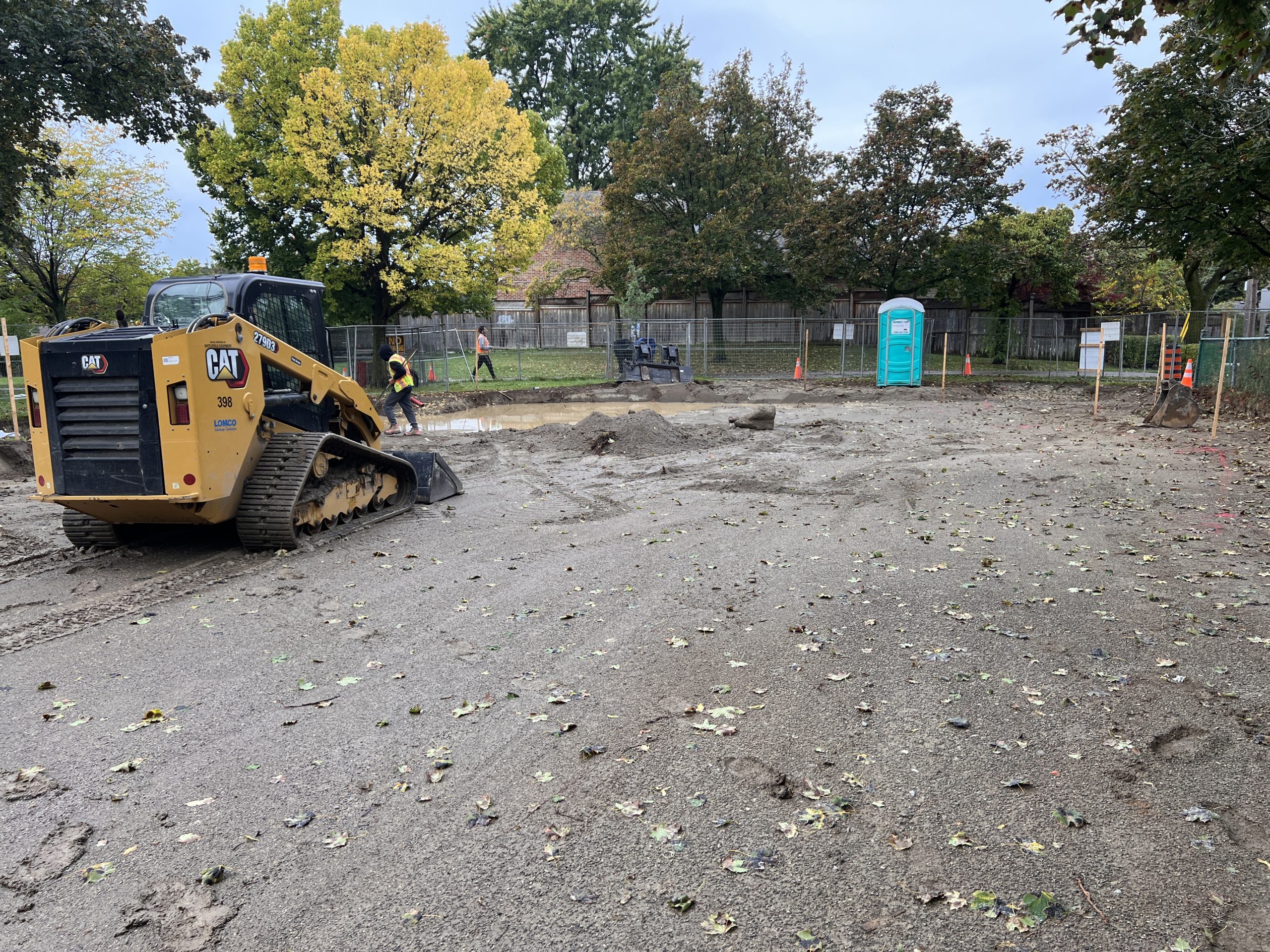 A yellow Cat compact track loader parked on a cleared construction site, with a worker in a high-visibility vest nearby. The site shows levelled dirt and some standing water, with trees and a fenced perimeter in the background.