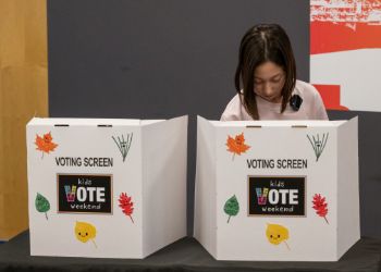 A child voting behind a voting screen that reads Kids Vote Weekend.