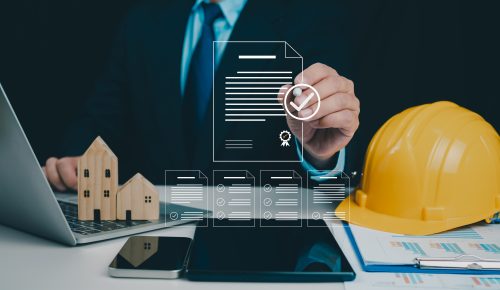 A person is writing on a semi-transparent document floating above a tablet, with a hard hat and model wooden buildings on the table as well on top of papers.