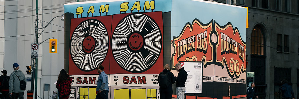 Large cube in downtown intersection with illustrations on the sides. Left visible side shows Sam the Record Man and right visible side shows Honest Eds. Some people walk in front.