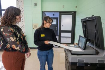 Election official assisting a voter with inserting a ballot into a vote-counting machine.